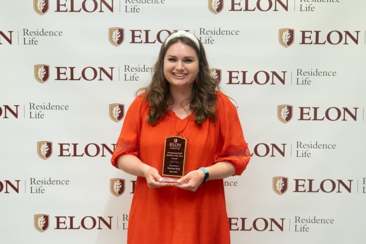 A smiling student in an orange dress holds a leadership award plaque in front of an Elon Residence Life backdrop.