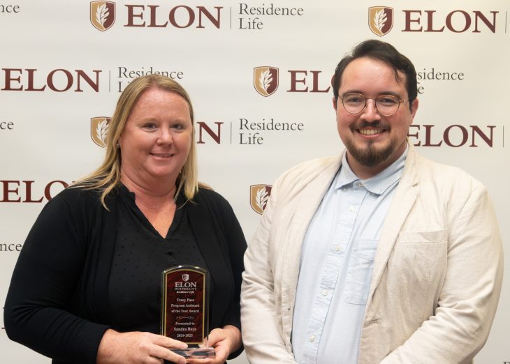 A woman holding a leadership award plaque stands beside a smiling man in front of an Elon Residence Life backdrop.