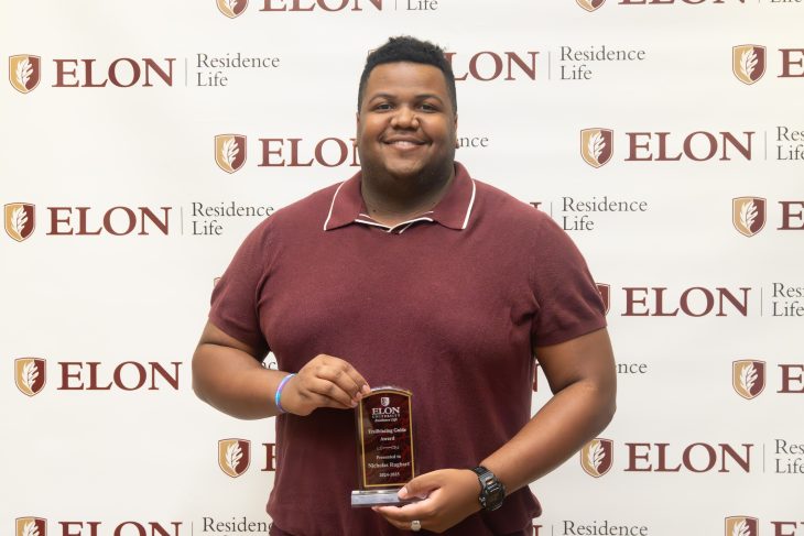 A smiling student in a maroon shirt holds a leadership award plaque while standing in front of an Elon Residence Life backdrop.