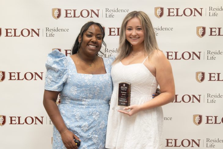 Two women smile in front of an Elon Residence Life backdrop, with one proudly holding a plaque recognizing her for a Residence Life leadership award.