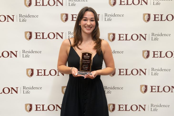 A smiling student in a black dress holds a leadership award plaque in front of an Elon Residence Life backdrop.