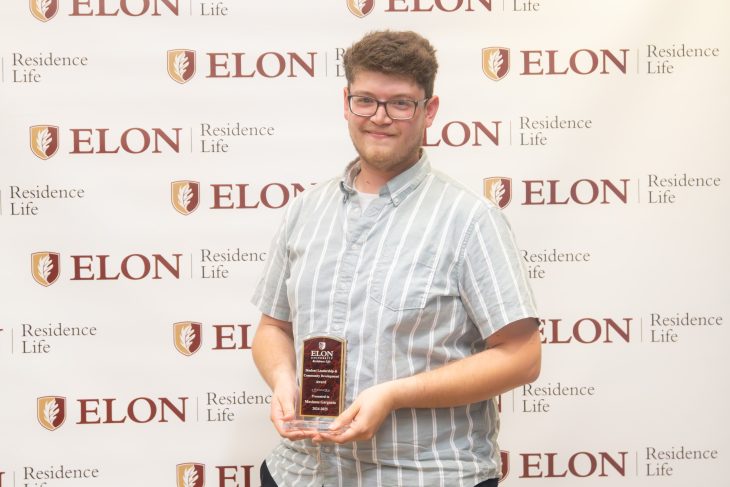 A student wearing glasses smiles while holding a leadership award plaque in front of an Elon Residence Life backdrop.