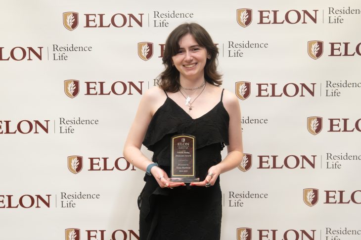 A student stands in front of an Elon Residence Life backdrop, holding a plaque and smiling proudly after receiving a leadership award.