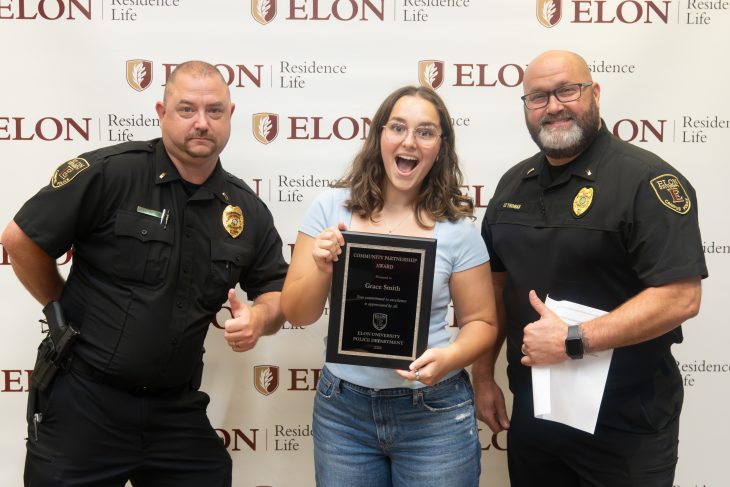 A student excitedly holds a Community Partnership Award plaque while standing between two smiling campus police officers giving thumbs up in front of an Elon Residence Life backdrop.