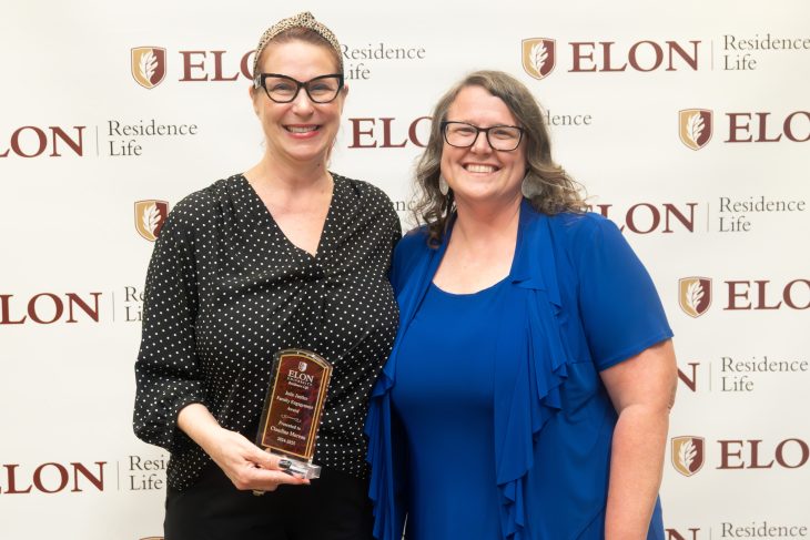 Two women smile in front of an Elon Residence Life backdrop, with one proudly holding a leadership award plaque.