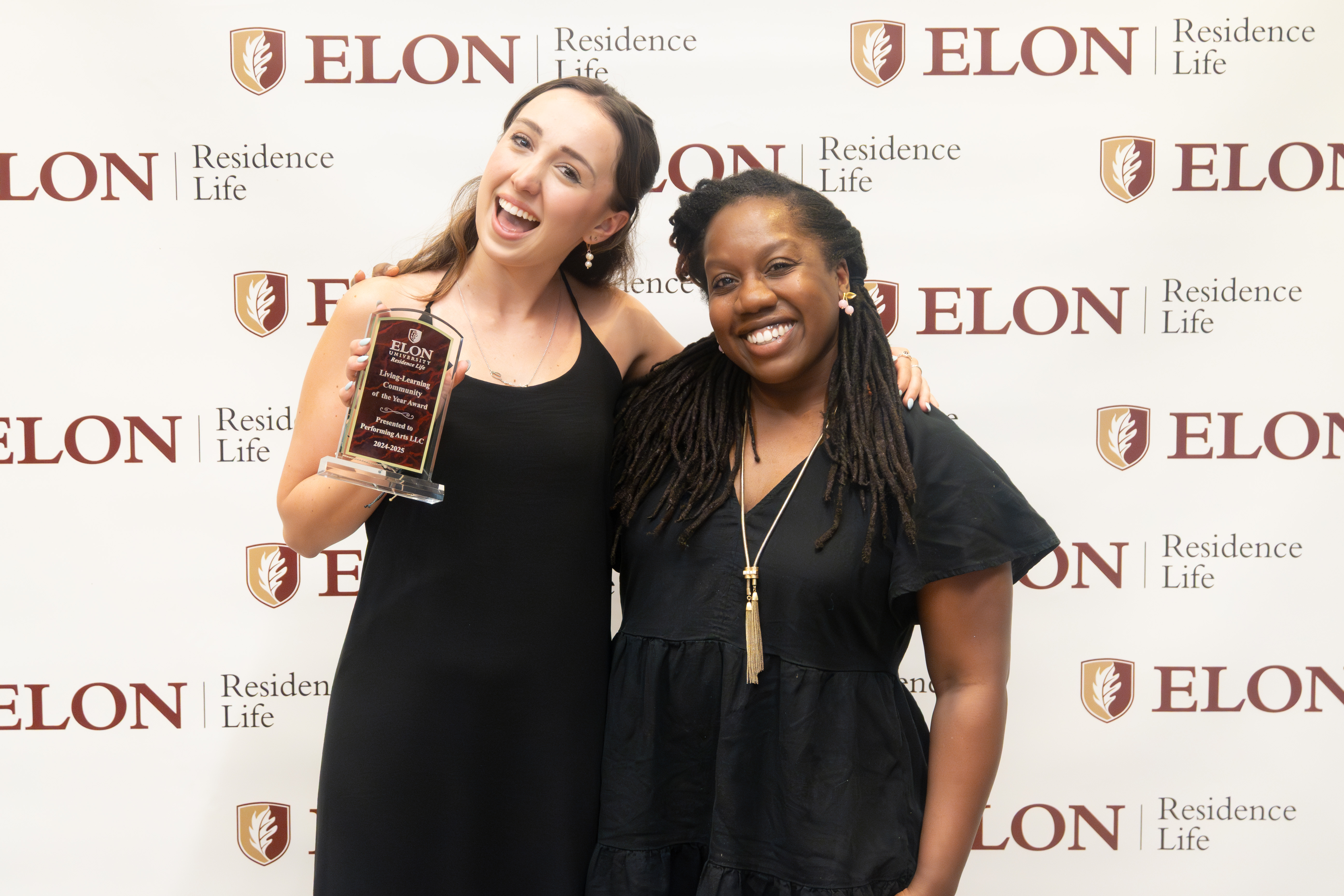 Two smiling women pose in front of an Elon Residence Life backdrop, one proudly holding a leadership award plaque while the other has her arm around her.