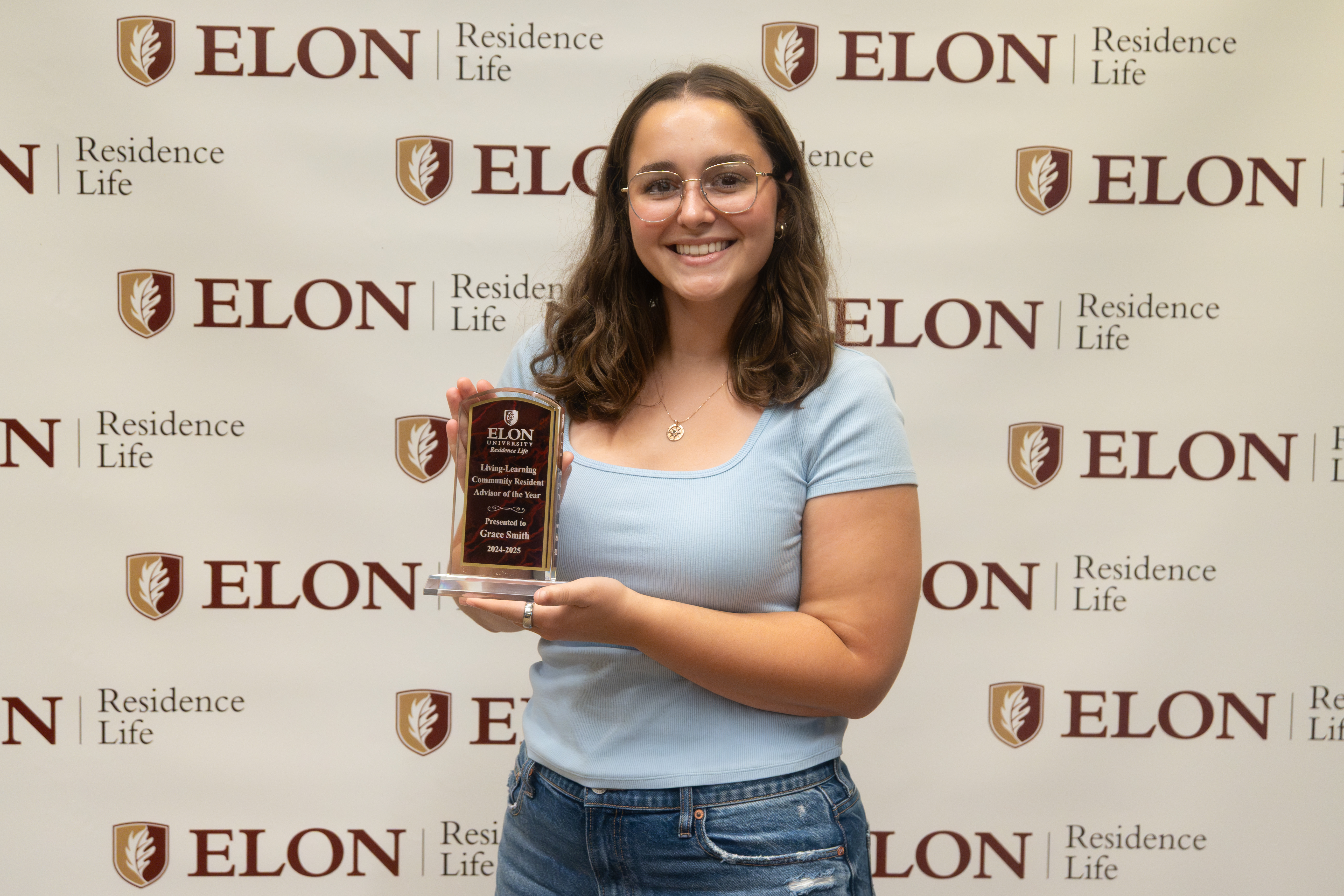A student stands in front of an Elon Residence Life backdrop, holding a plaque and smiling proudly after receiving a award.