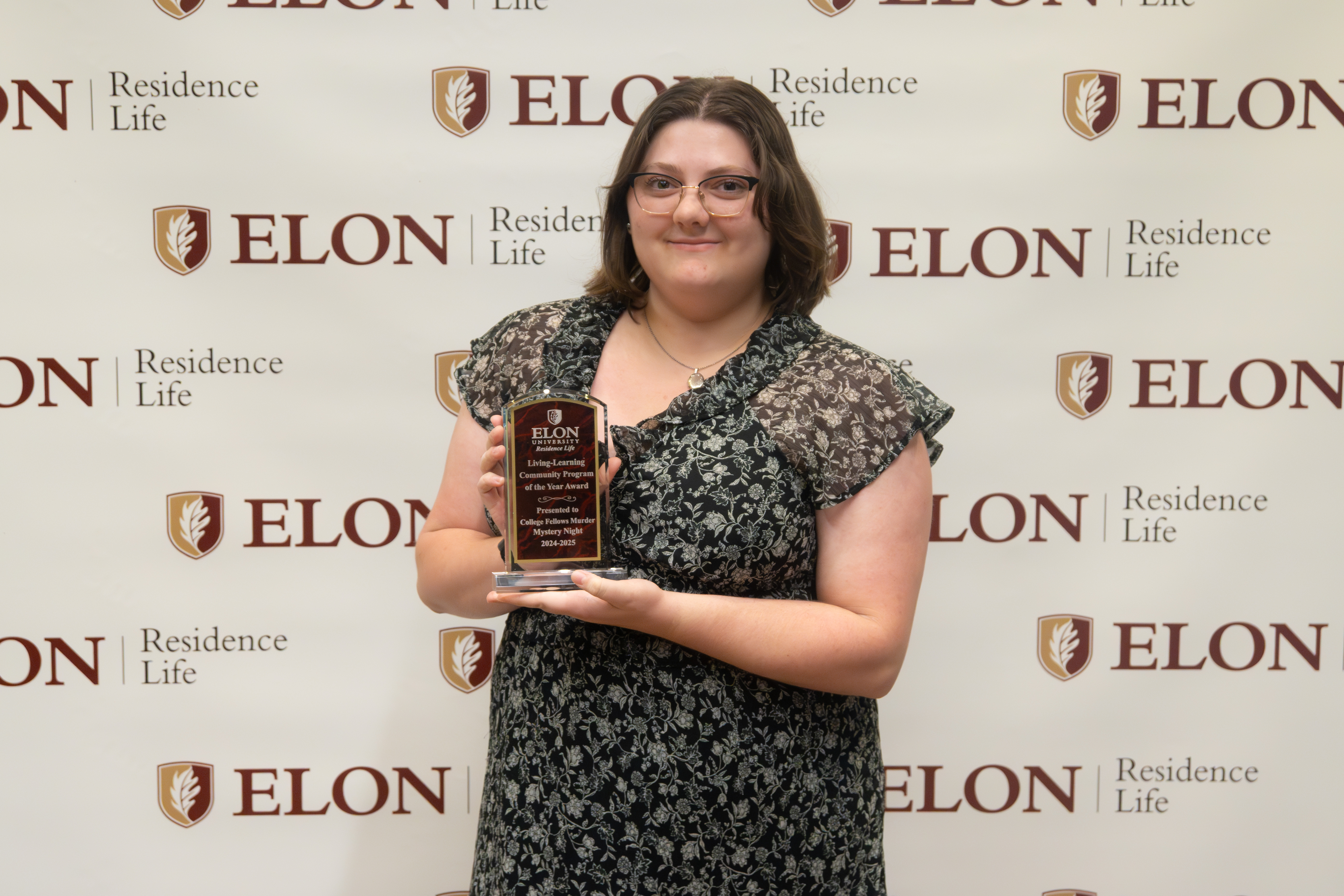 A student stands in front of an Elon Residence Life backdrop, holding a plaque and smiling proudly after receiving a leadership award.