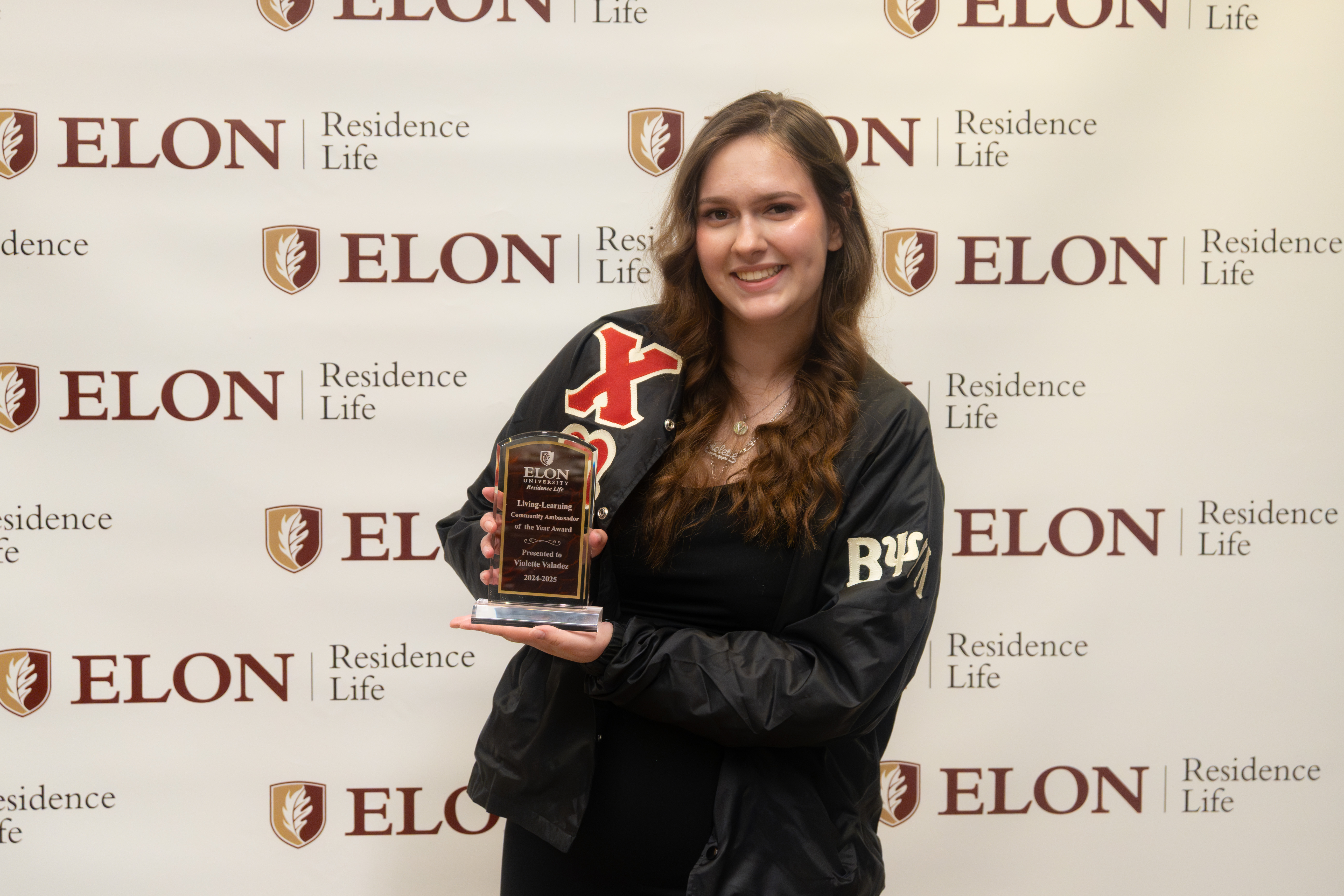 A smiling student stands in front of an Elon Residence Life backdrop, holding a plaque that recognizes their achievement in leadership.