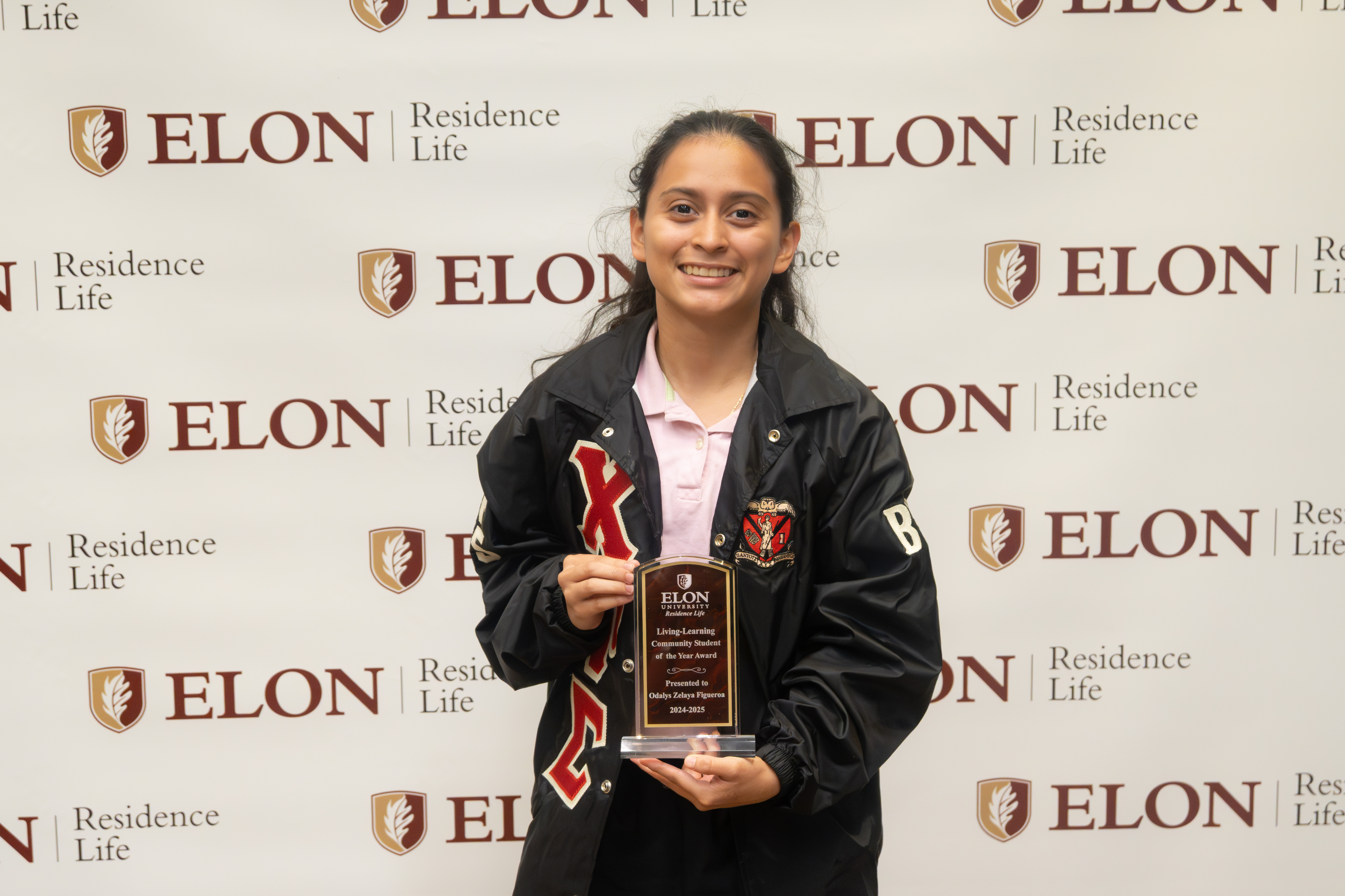 A smiling student stands in front of an Elon Residence Life backdrop, holding a plaque that recognizes their achievement in leadership.