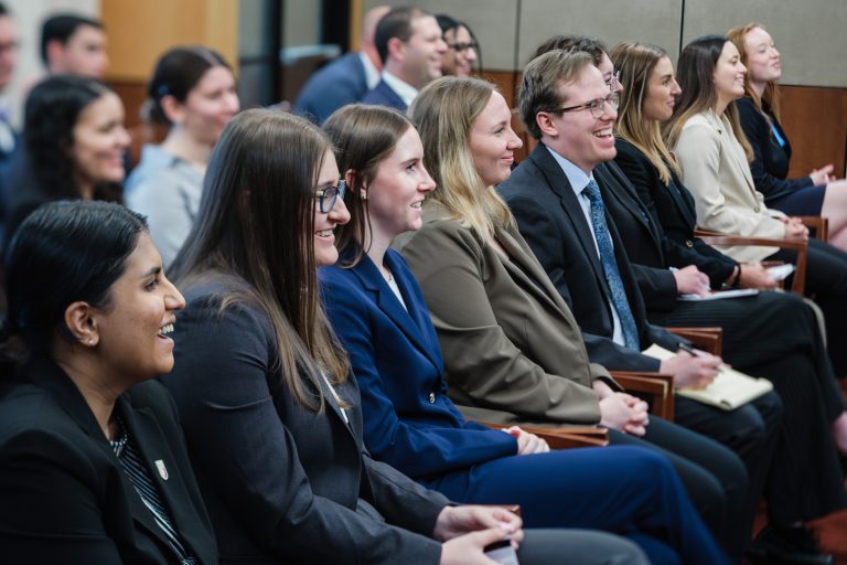 Several rows of students dressed professionally, seated in a courtroom. They are smiling and laughing.