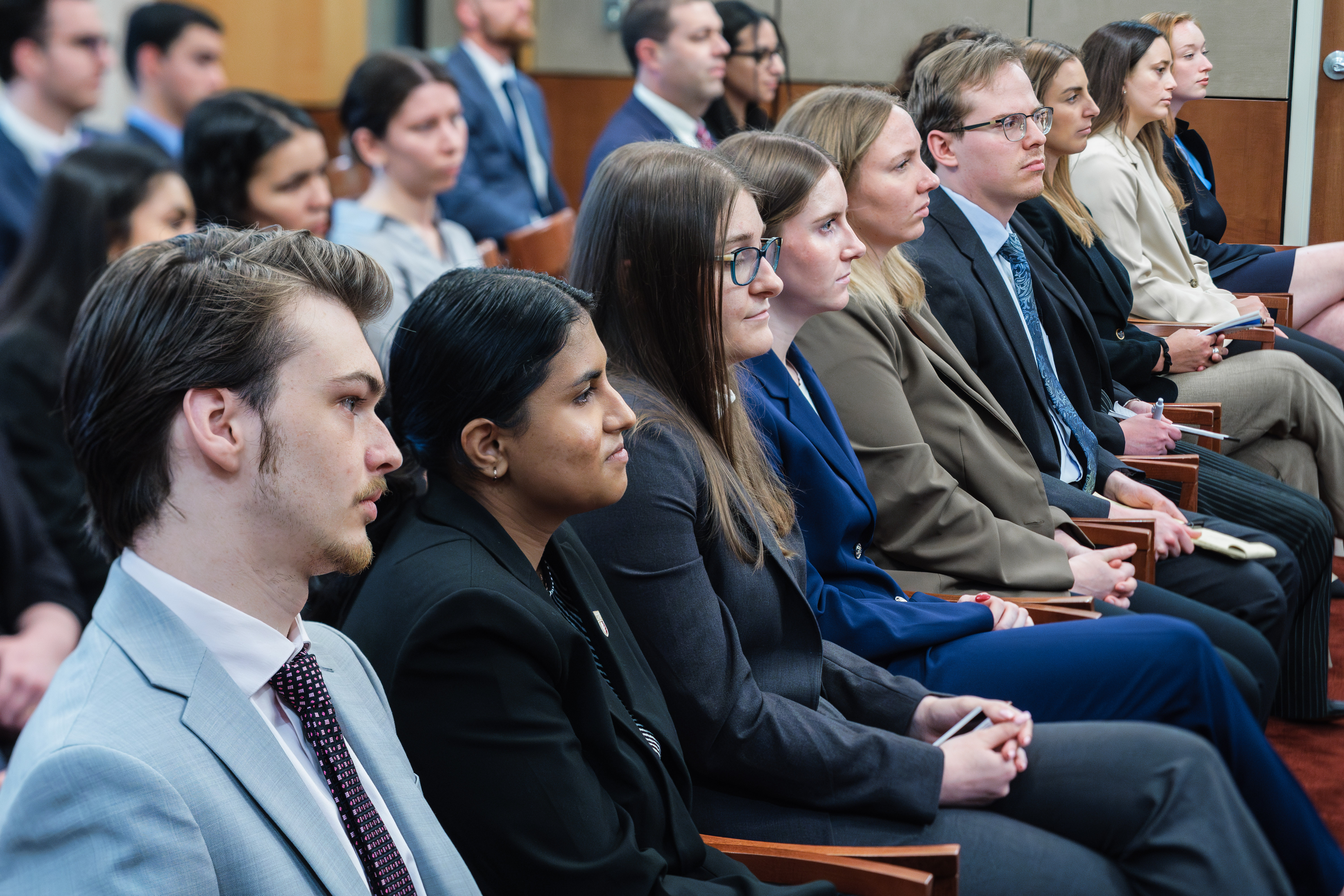 Several rows of students seated in a courtroom. They are dressed professionally and listening intently.