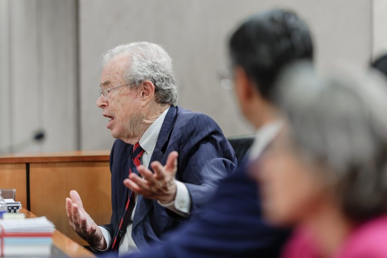 A man gestures emphatically speaking into a microphone. Two subjects are seen in the foreground listening to him.