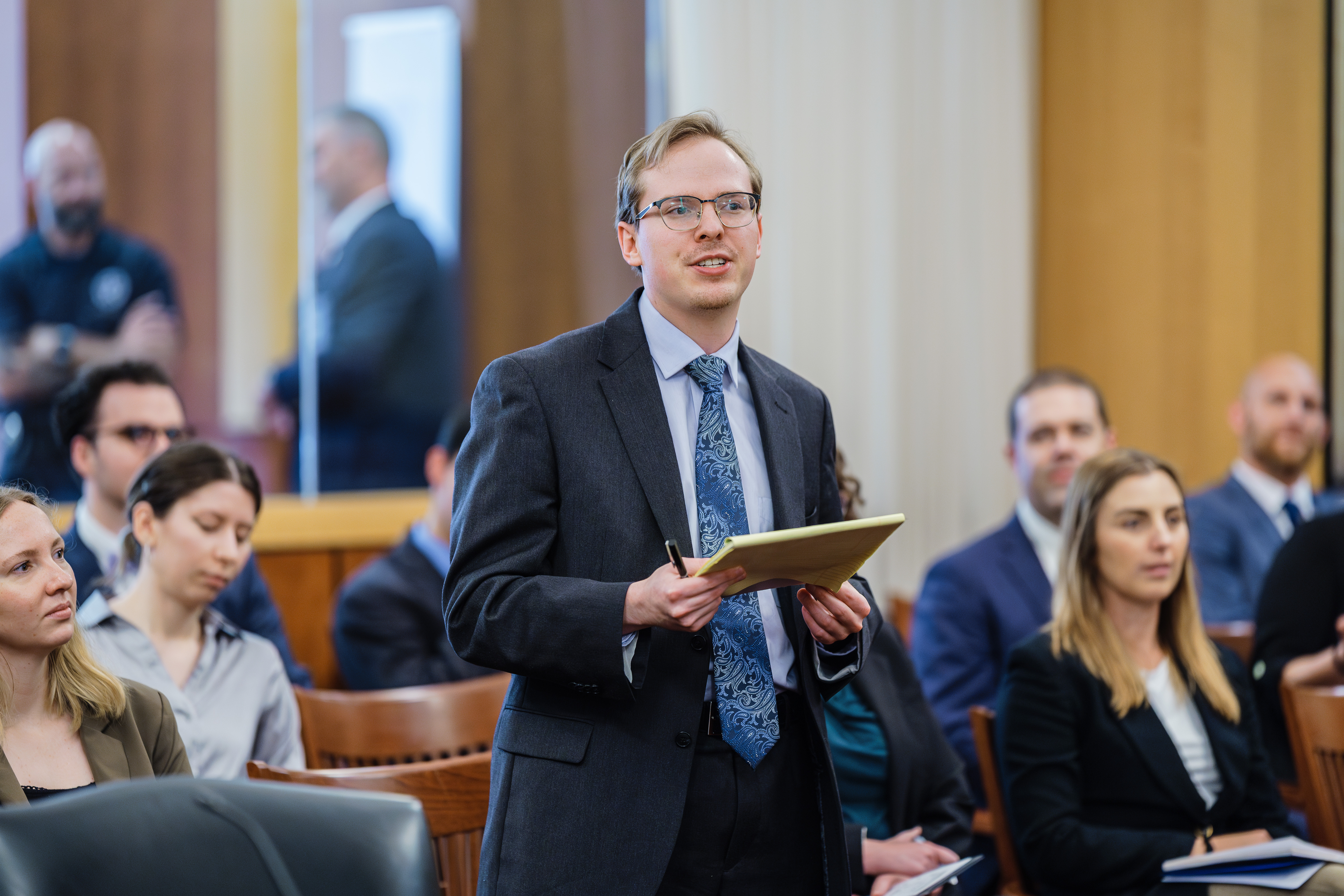 A young man wearing a suit and holding a legal pad stands before a crowded courtroom.