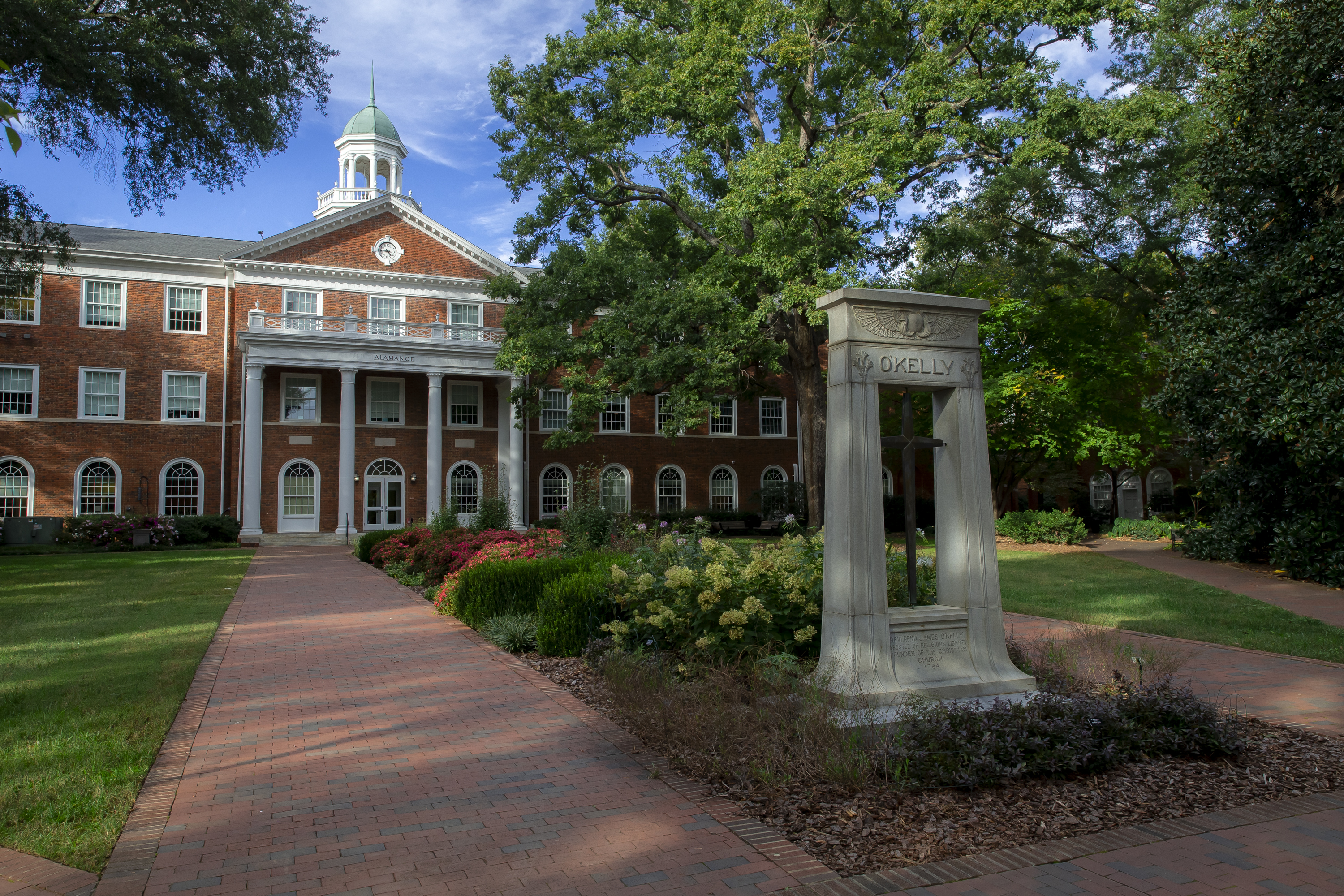 A view of Alamance Building at Elon University on a bright day, framed by vibrant landscaping and mature trees. The iconic O’Kelly monument with a bronze bell and the name 