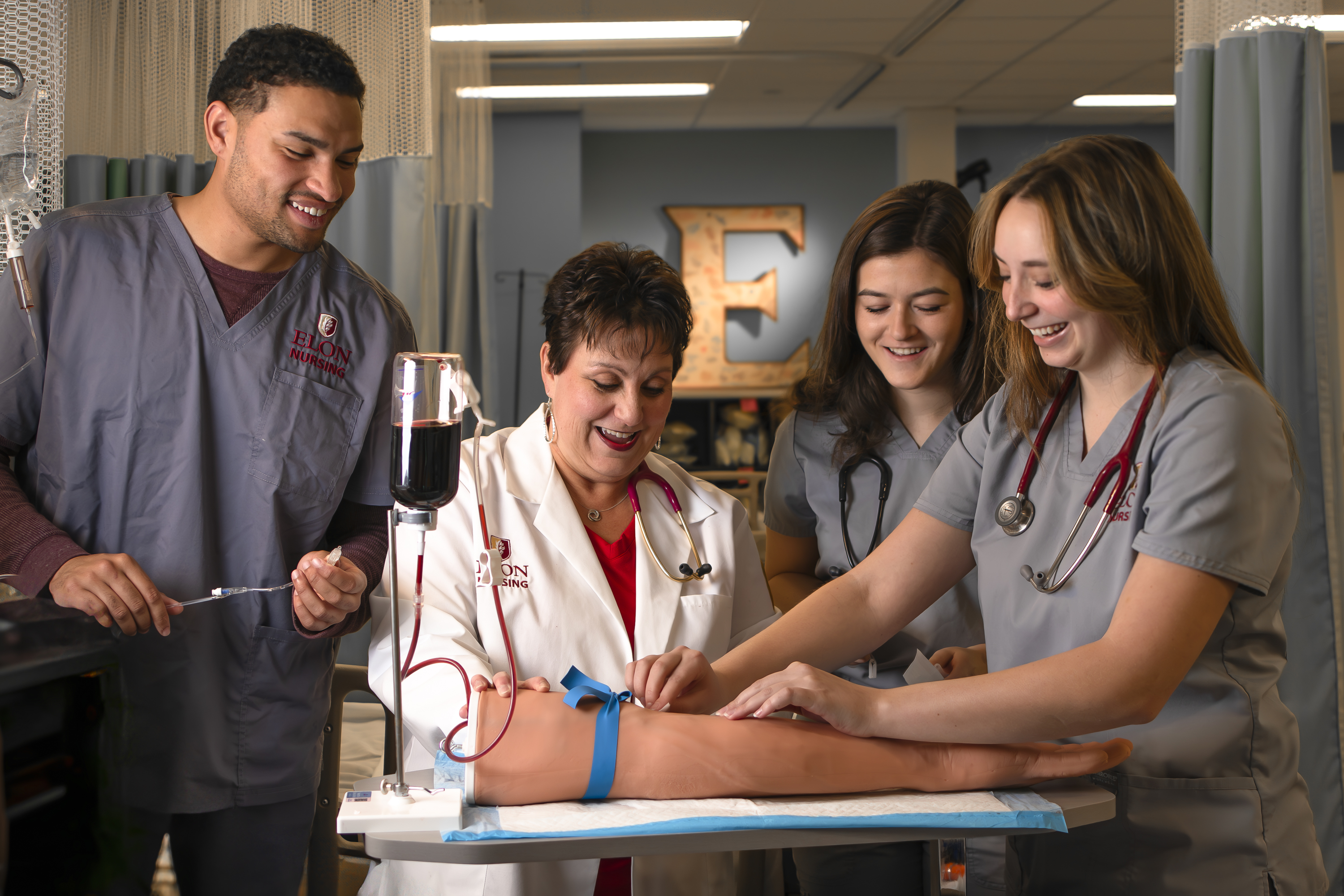 A group of nursing students and an instructor smile and collaborate while practicing on medical training mannequin arm.