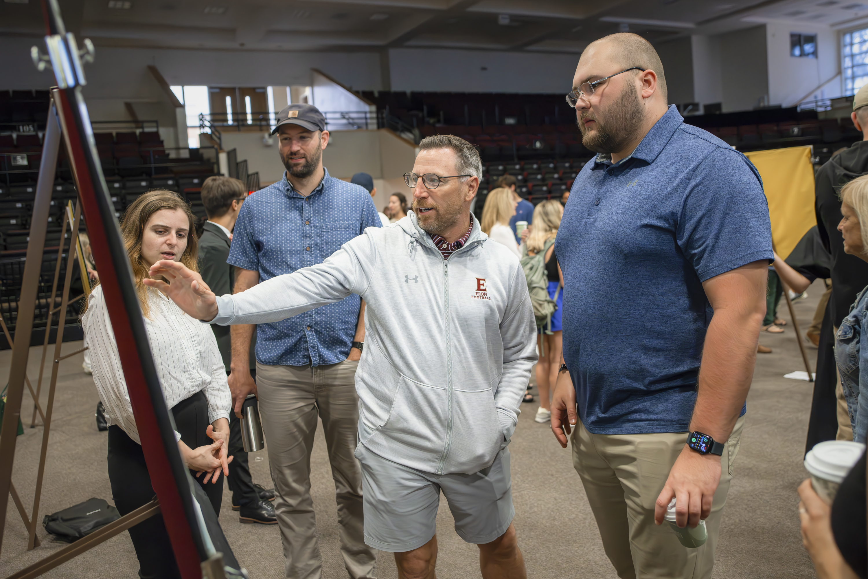 A group of people stands around a presentation board as a man in an Elon University hoodie gestures while explaining something during an indoor academic or professional event.