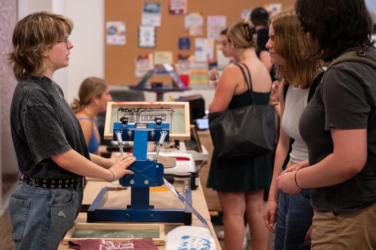 A student demonstrates screen printing techniques to a group of curious onlookers at a maker fair booth.