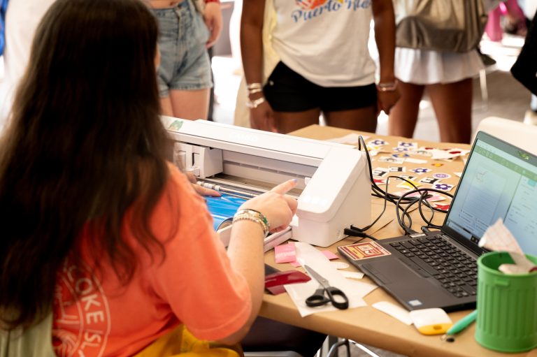 A student operates a vinyl cutting machine at a craft table covered in stickers, tools, and a laptop, while others watch from across the table.