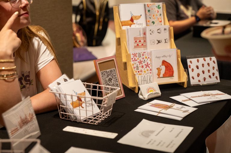 A vendor sits behind a table showcasing a variety of whimsical, illustrated greeting cards arranged in neat displays.