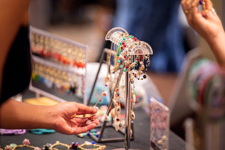 Handmade jewelry, including colorful beaded bracelets and necklaces, hangs neatly on a display rack as a hand reaches to pick one.