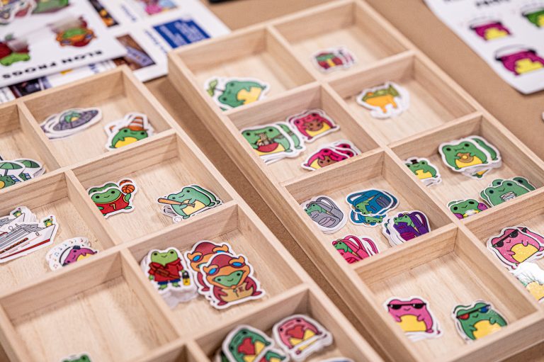 Wooden trays display dozens of playful frog-themed stickers in various poses and outfits at a vendor’s table.