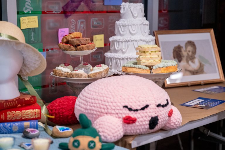 A colorful display table features plush toys, painted rocks, faux desserts, a layered white cake, and vintage books under a soft light.