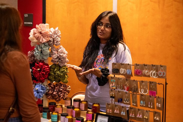 A woman stands behind a display of handmade earrings, scrunchies, and candles, speaking to a customer at a craft or vendor fair.