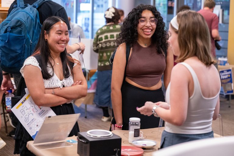 Two students smile and chat with a vendor at a table filled with crafting supplies and a demonstration setup.