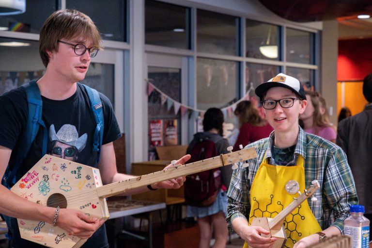 Two students show off and play handmade wooden instruments at a bustling indoor event.