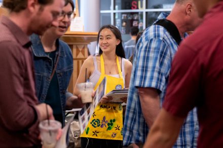 A smiling student in a yellow apron holds a brochure and engages with attendees at a bustling event.