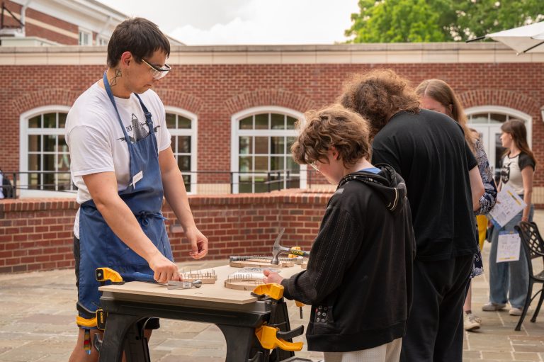 A person in a blue apron guides a child through a hands-on woodworking activity at an outdoor table.