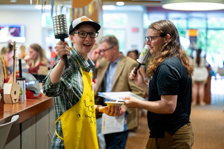 A cheerful student in a yellow apron enthusiastically holds up a metal percussion instrument while talking with another attendee holding a lollipop-shaped item.