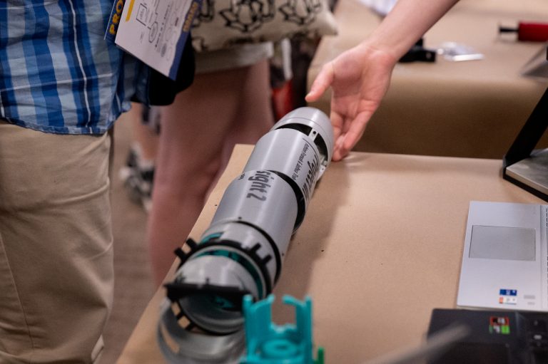A hand reaches toward a cylindrical prototype labeled “SignalZ” displayed on a table among other projects.