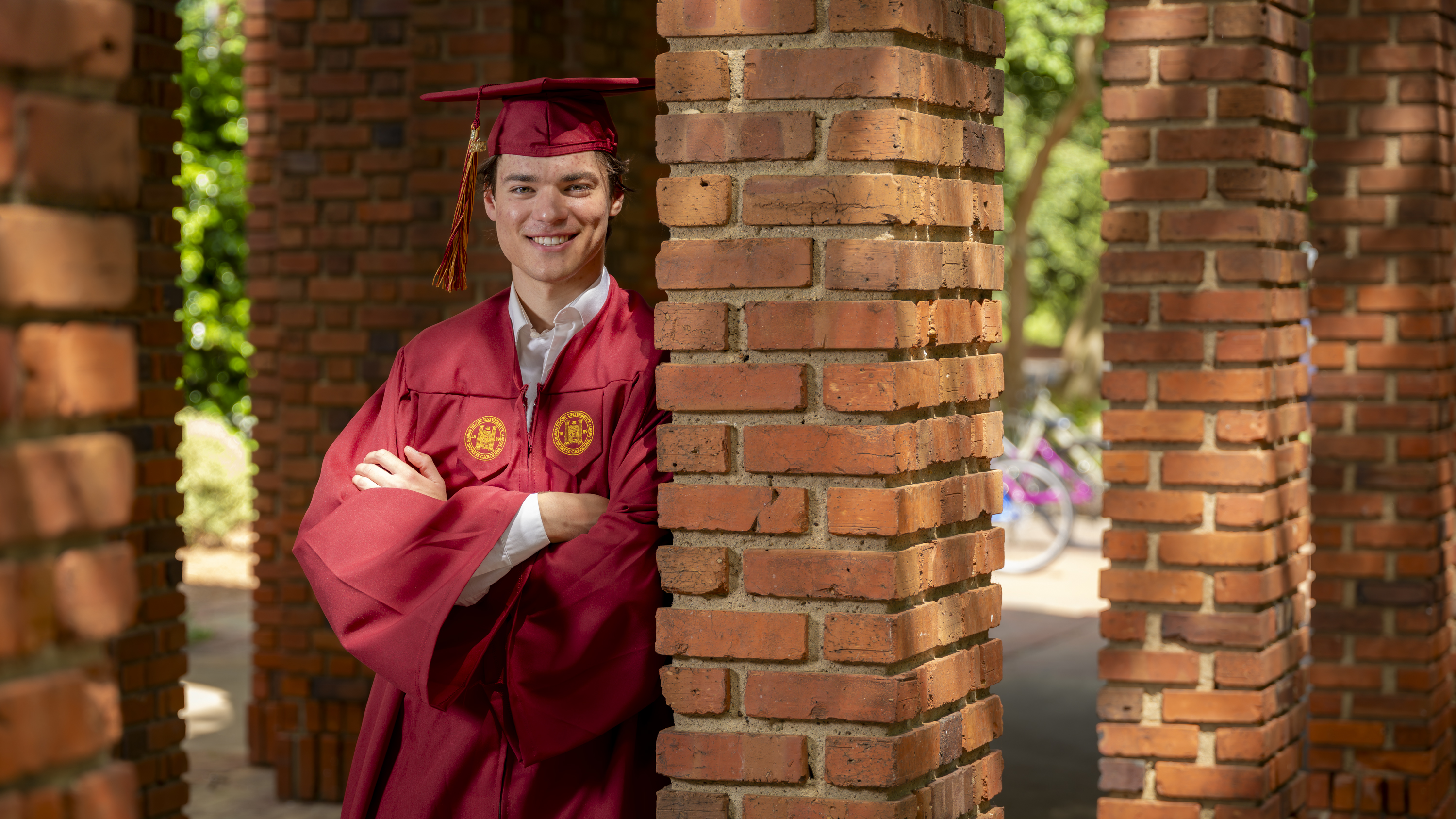 A graduate in a maroon cap and gown smiles with arms crossed while leaning against a brick column.