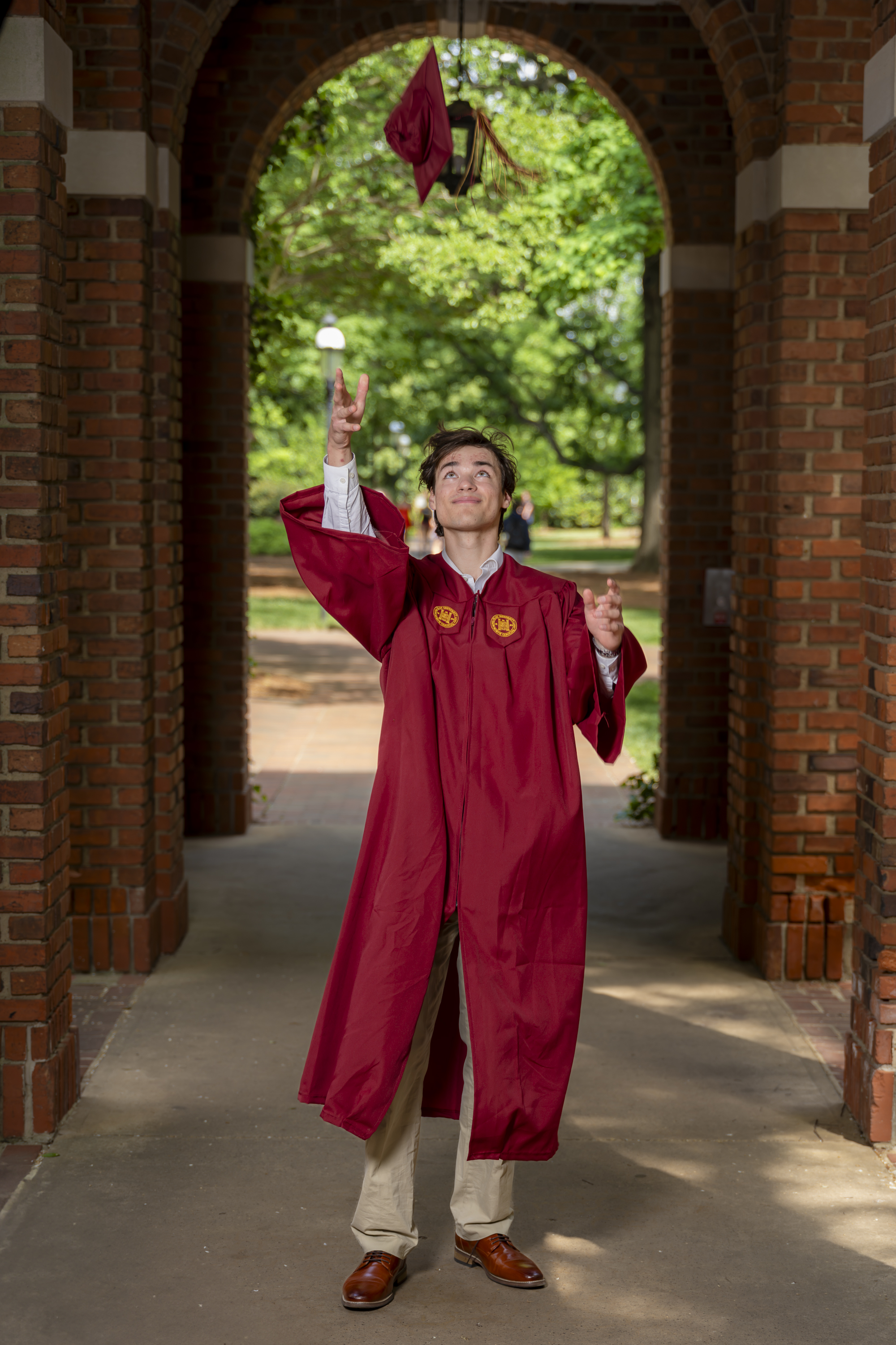A graduate in a maroon gown throws his cap into the air under a brick archway.