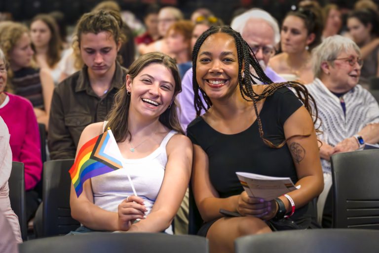Two audience members smile for the camera, one holding a small rainbow flag, surrounded by a diverse and engaged crowd.