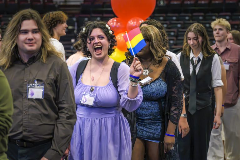 A group of students walk in a celebratory procession, with one student in a purple dress excitedly waving a pansexual pride flag.