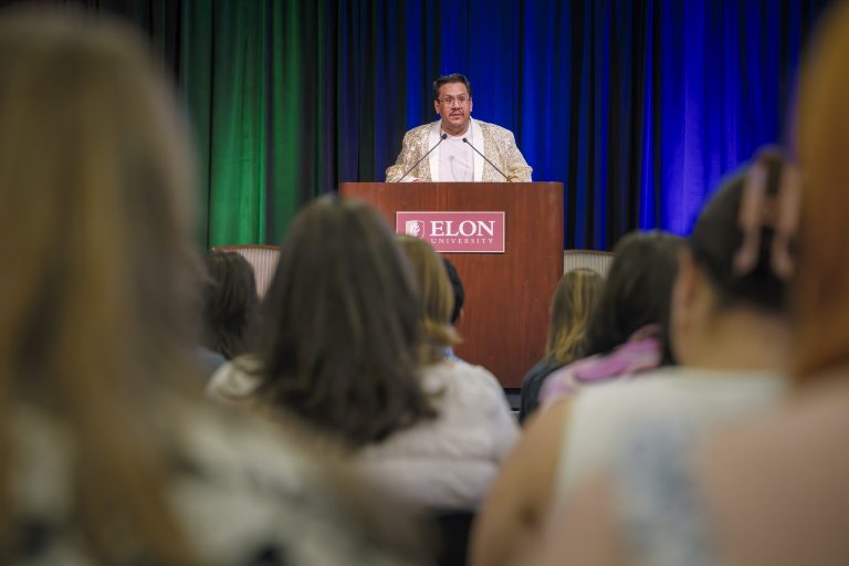 A speaker stands at a podium with the Elon University logo, addressing an audience seated in front of a backdrop lit with green, blue, and purple lights.