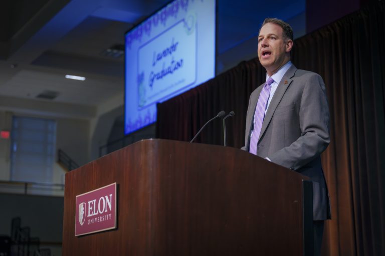 A speaker in a suit stands at the Elon University podium addressing the audience during a Lavender Graduation ceremony.