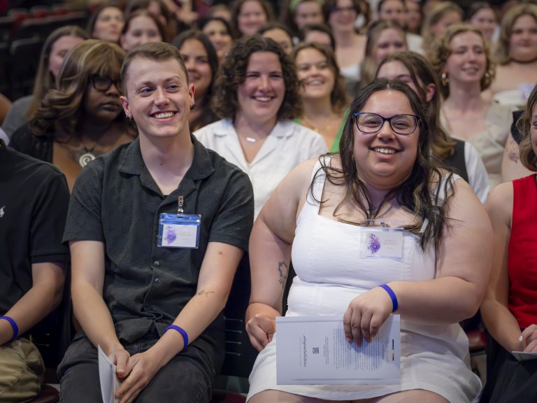Two smiling students sit together in the front row of a large, cheerful audience at an Elon University event.