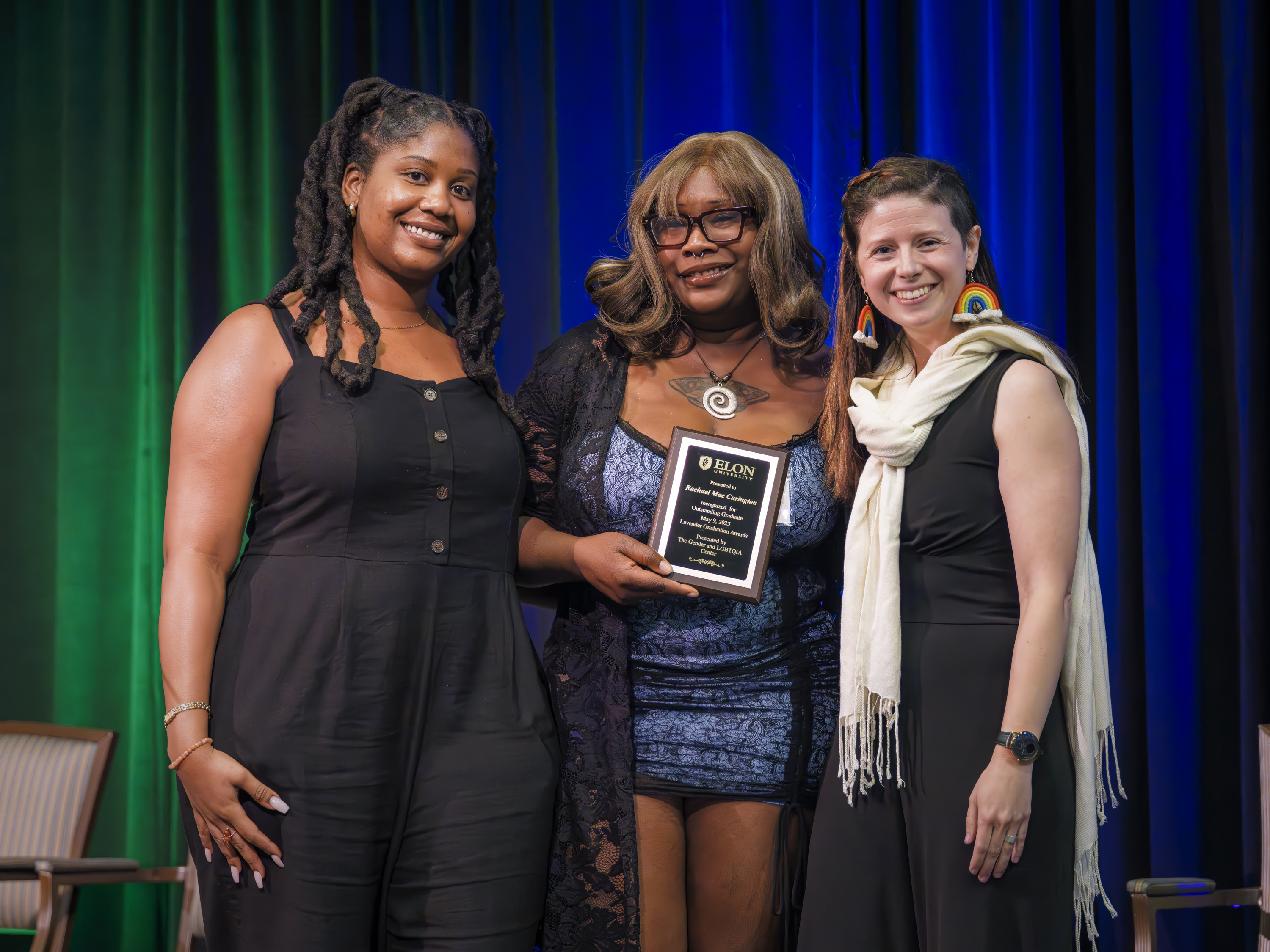 Three women smile on stage as the woman in the center holds an award plaque at an Elon University event.