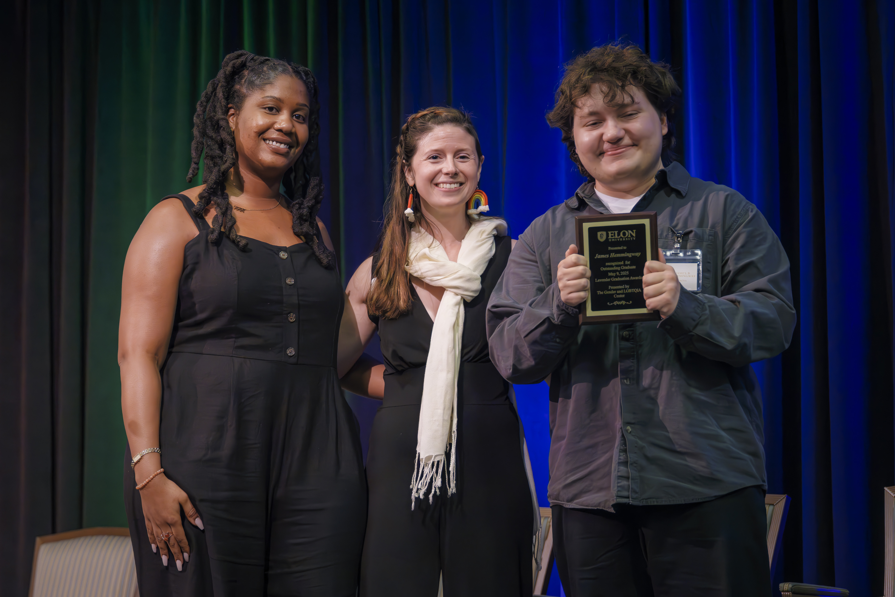 Three people smile on stage as the person on the right holds an award plaque at an Elon University event.