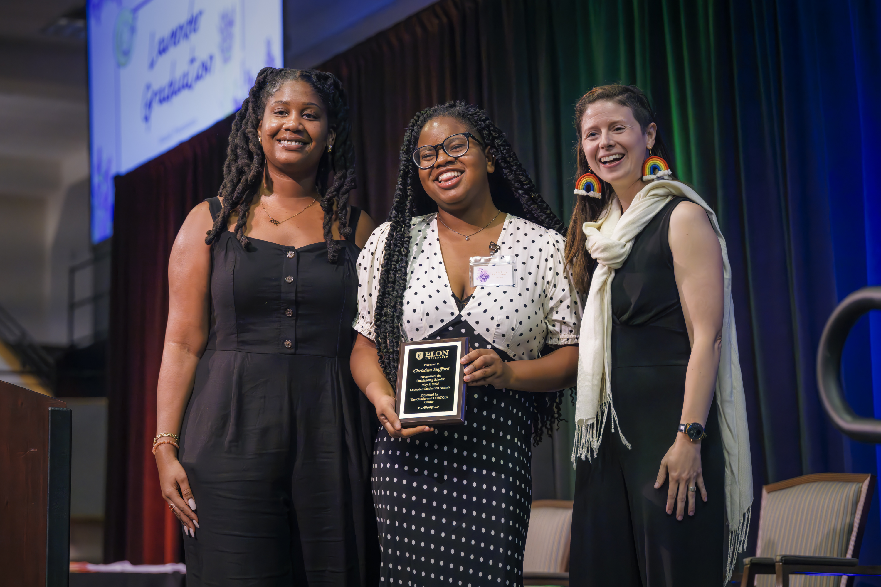 Three women smile on stage as the woman in the center holds an award plaque at an Elon University event.
