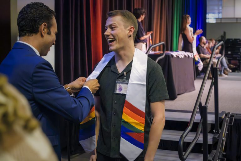 A smiling student wearing a rainbow graduation stole is helped by a man in a suit during a ceremony at Elon University.