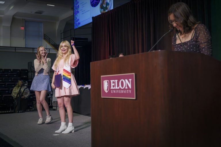 A student in a pink dress and rainbow stole raises a fist in celebration on stage while another claps nearby, as a speaker reads from the podium at an Elon University event.