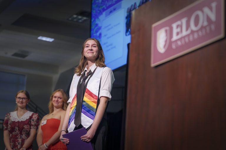 A student wearing a rainbow stole and black tie smiles confidently on stage beside the Elon University podium as two people stand behind them.