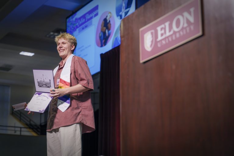 A student grins while holding up a certificate and folder on stage, wearing a rainbow stole near the Elon University podium.