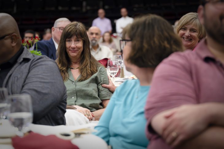 A woman sits smiling at a banquet table, surrounded by colleagues, as another person places a supportive hand on her arm during an awards ceremony.