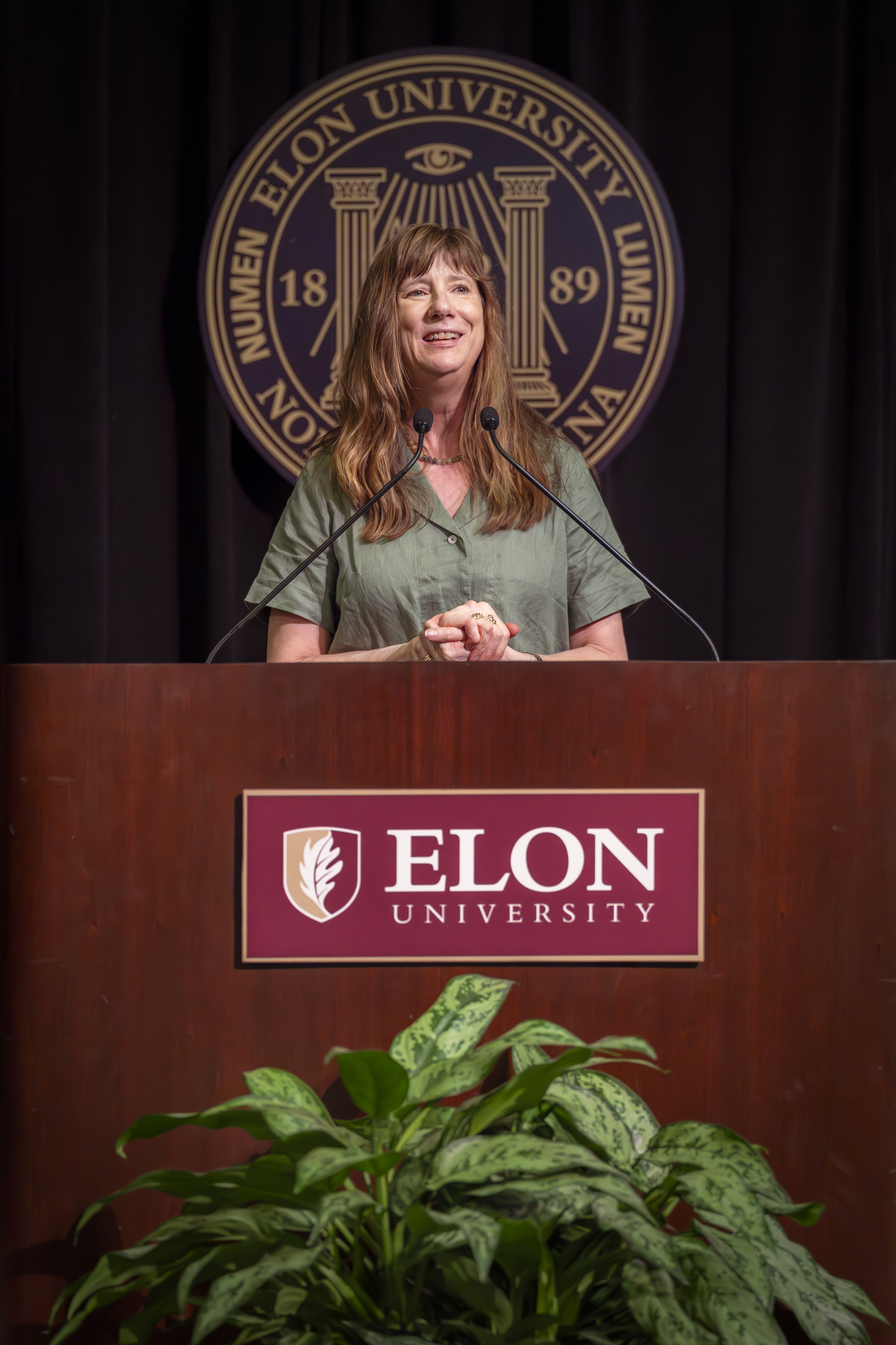 A woman smiles as she speaks at a podium bearing the Elon University logo, with the university seal displayed behind her.